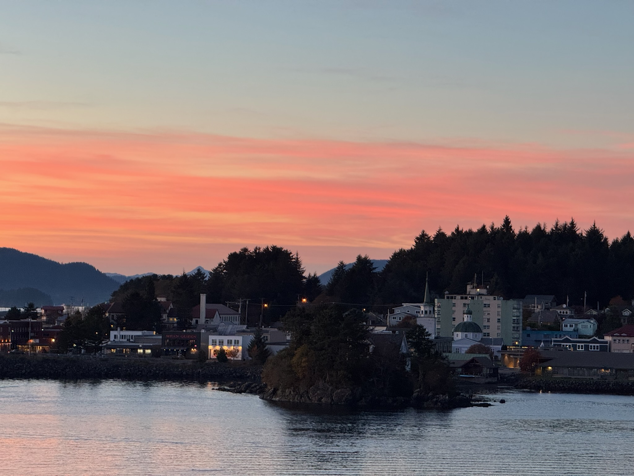 Alaskan waterfront town at dusk with pink-orange sky, mountains, church steeple, and illuminated buildings