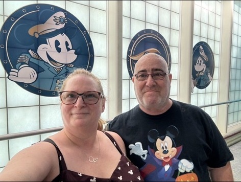 Lawrence and his wife in the Disney Fantasy concierge boarding area at Port Canaveral, with Mickey Mouse nautical panels behind them