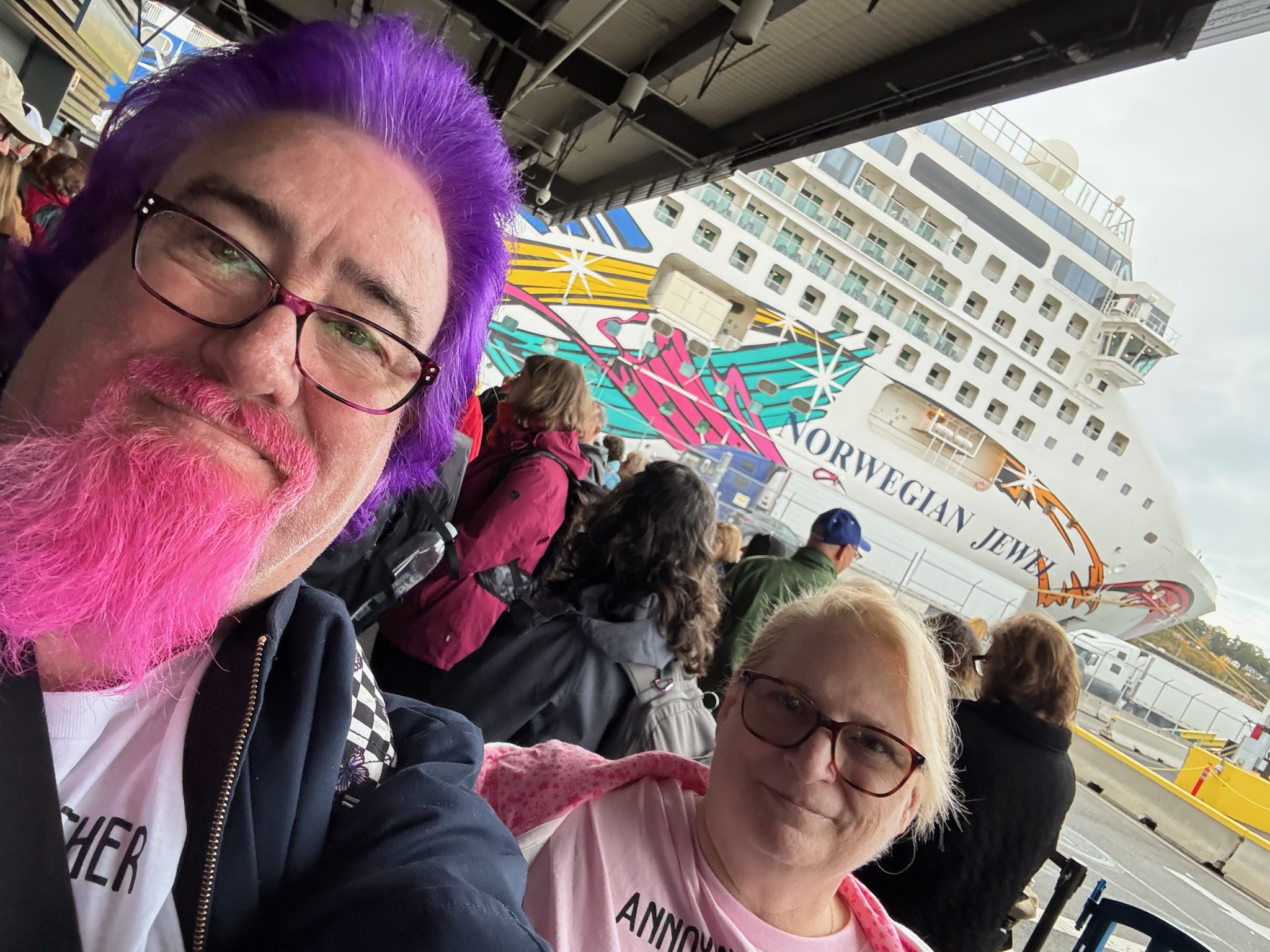 Lawrence and his wife smiling in the boarding queue in front of the colorful Norwegian Jewel cruise ship